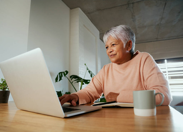 Elderly multi-ethnic female sitting at home counter scrolling on her laptop, smiling. - Φωτογραφία, εικόνα