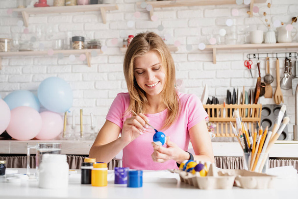 Happy Easter. Young millennial woman in pink shirt coloring easter eggs in the kitchen - Foto, imagen