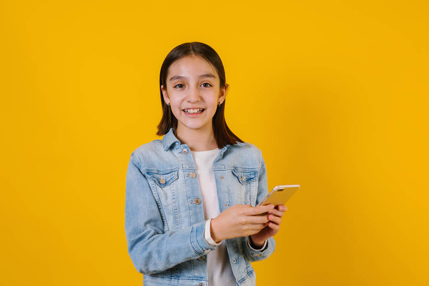 Latin child girl on holding a mobile phone on yellow background in Mexico Latin America - Photo, Image