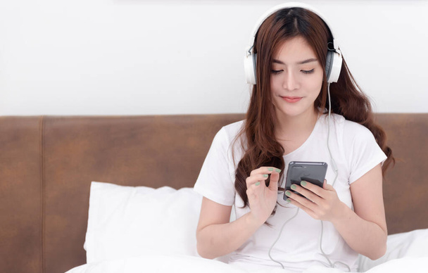 Beautiful Asian women wearing headphones listening to music streaming from their smartphones. On the bed in the white bedroom - Photo, Image