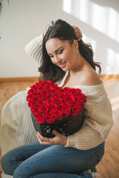 Όμορφη νεαρή μελαχρινή γυναίκα σε πλεκτή ζακέτα κρατά ένα κουτί δώρου σε σχήμα καρδιάς με ένα μπουκέτο κόκκινα τριαντάφυλλα στα χέρια της. Ημέρα του Αγίου Βαλεντίνου. - Φωτογραφία, εικόνα