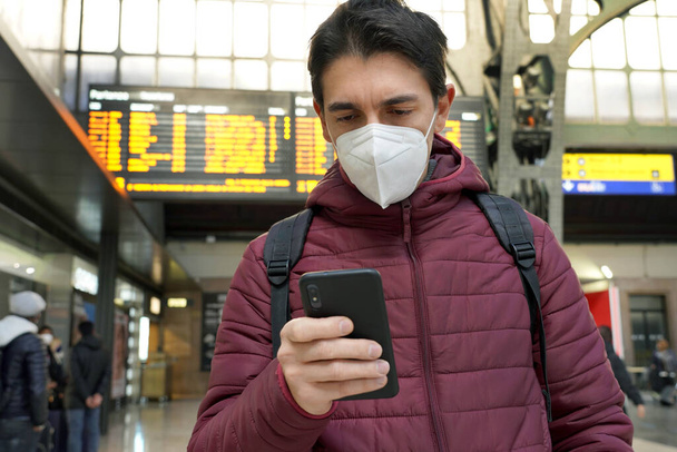 Voyageur homme portant le masque protecteur KN95 FFP2 à l'aéroport. Jeune homme caucasien avec derrière les horaires tableau des départs arrivées vérifier billet en ligne de son vol. - Photo, image