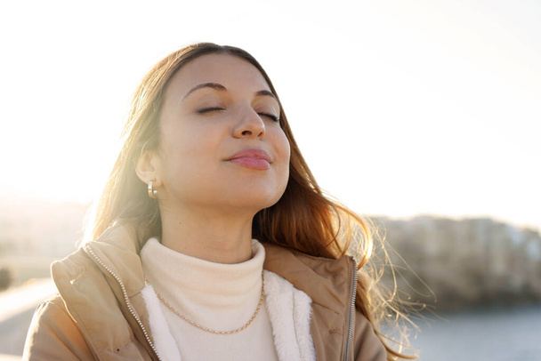Молода чарівна жінка дихає свіжим повітрям, розслабляючись і посміхаючись із закритими очима на заході сонця
 - Фото, зображення