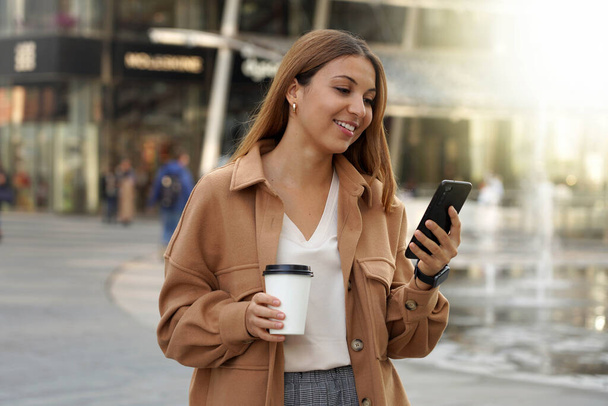 Entrepreneur young. Busy woman. Smartphone app. Office worker outdoors. Cappuccino coffee cup. Shopping mall. Architecture modern. Urban background. - Foto, immagini
