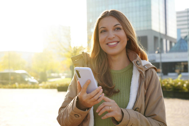 Усміхнена красива дівчина тримає смартфон зовні на заході сонця
 - Фото, зображення