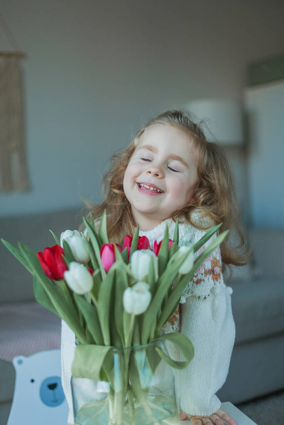 Cute little girl of 3 years old in a cozy knitted sweater hugs a vase with a bouquet of white and pink tulips in a home interior. Home comfort. - Foto, Bild