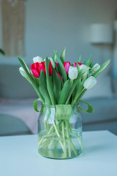 春のインテリアで白いコーヒーテーブルの上に大きなガラス花瓶にチューリップの美しい多色の花束。居心地の良い家。春. - 写真・画像