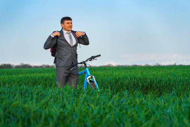 hombre de negocios se pone una mochila, posa con bicicleta en un campo de hierba verde, vestido con un traje de negocios, hermosa naturaleza en primavera, concepto de negocio - Foto, Imagen