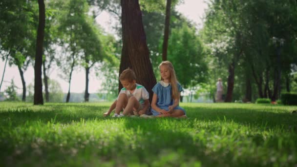 Laughing kids sitting under tree in green park. Happy active childhood concept. - Footage, Video