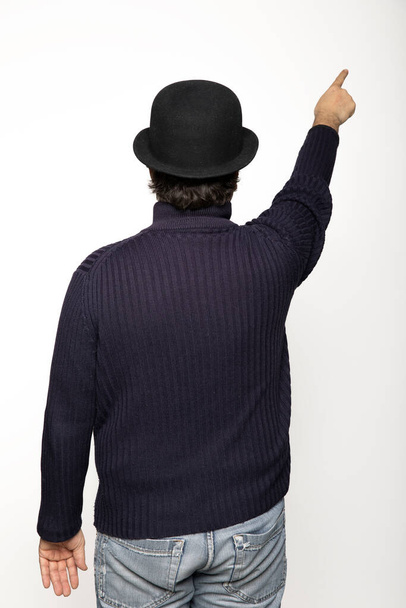 Man turned from behind indicates with his finger an upward point. He is wearing a blue sweater, jeans and a bowler hat. Isolated on white background - Photo, image