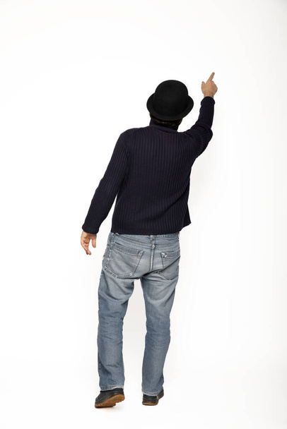 Man from behind indicates with his finger an upward point while walking. He is wearing a blue sweater, jeans and a bowler hat. Isolated on white background - Foto, Bild