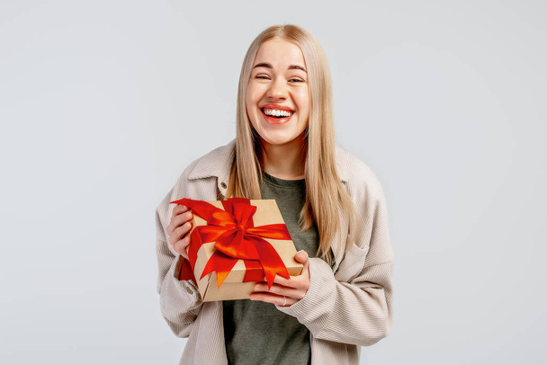 Smiling young blonde woman hold gift box with red ribbon, standing isolated on light background. New Year Women's Day birthday holiday concept - Foto, afbeelding