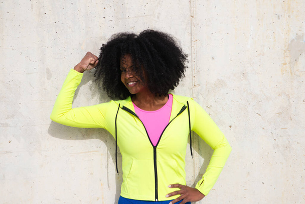 Schöne junge Afroamerikanerin in leuchtend grüner und pinkfarbener Sportbekleidung auf grauem Betonwandhintergrund. Die Frau drückt sich anders aus. Lachend, ernst, glücklich, traurig, denkend - Foto, Bild