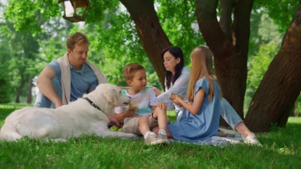 Gelukkige familie voeden witte hond op picknick. Mensen heve plezier op groen gras. - Video