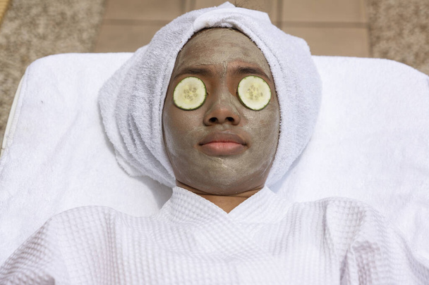 змішана раса чорна жінка лежить вершкова маска для обличчя і розслабляється і має масаж обличчя з майстром масажу обличчя, який носить маску для обличчя біля басейну СПА-курорту.
. - Фото, зображення