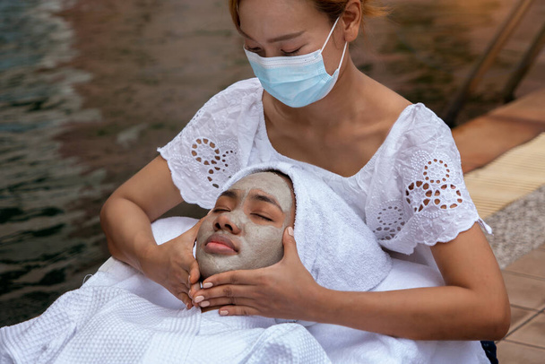 Afrikai amerikai nő fekvő krémes arc maszk és pihenjen, és egy arc masszázs egy arc masszázs technikus visel maszkot a medence spa üdülő.Wellness és szépség egészségügyi koncepció. - Fotó, kép