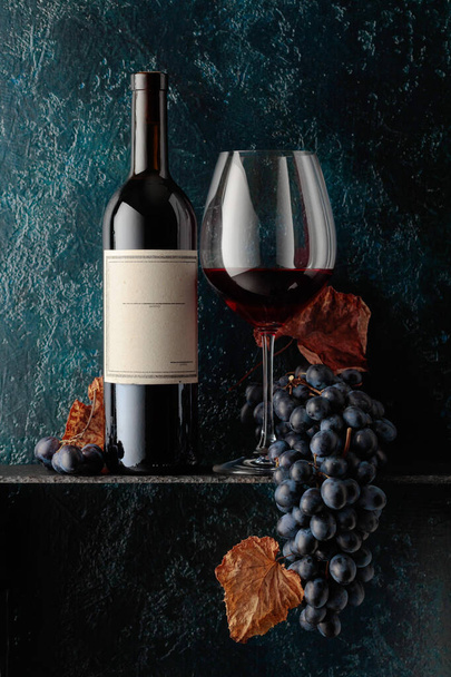 Red wine and blue grapes with dried up vine leaves on an old dark blue background. On a bottle old empty label. - Photo, Image