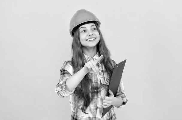 fröhliches Teenie-Mädchen in Helm und kariertem Hemd, das Notizen auf Klemmbrett macht, beschäftigt - Foto, Bild