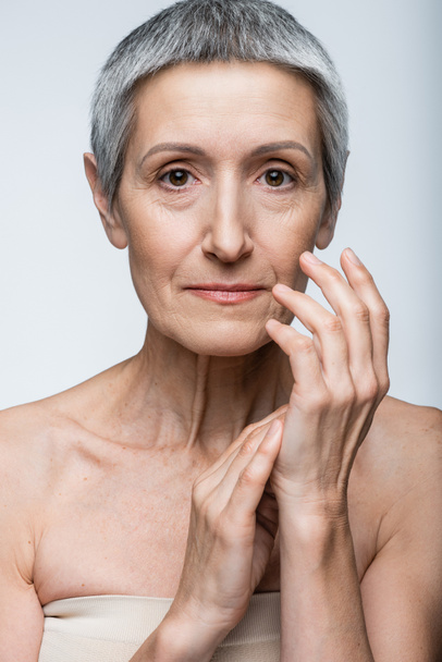 middle aged woman with bare shoulders touching hands isolated on grey - Foto, Imagen