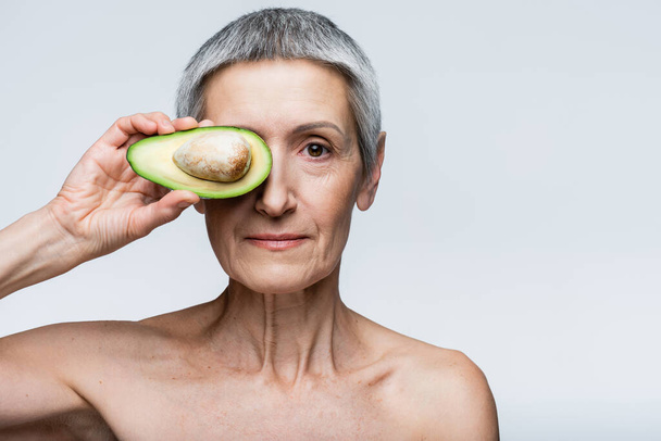 vrouw van middelbare leeftijd bedekken oog met de helft van avocado geïsoleerd op grijs - Foto, afbeelding