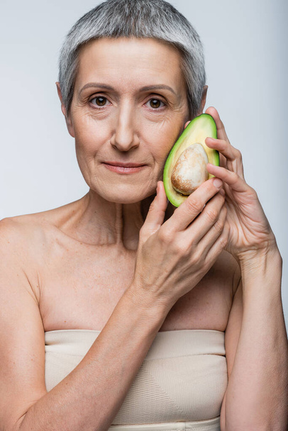 vrouw van middelbare leeftijd met de helft van de biologische avocado geïsoleerd op grijs - Foto, afbeelding