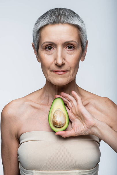 middle aged woman holding half of green and ripe avocado isolated on grey - Photo, Image