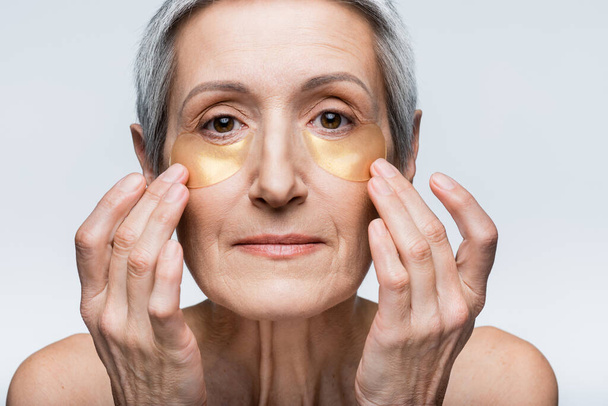 middle aged woman applying golden eye patches while looking at camera isolated on grey - Fotoğraf, Görsel