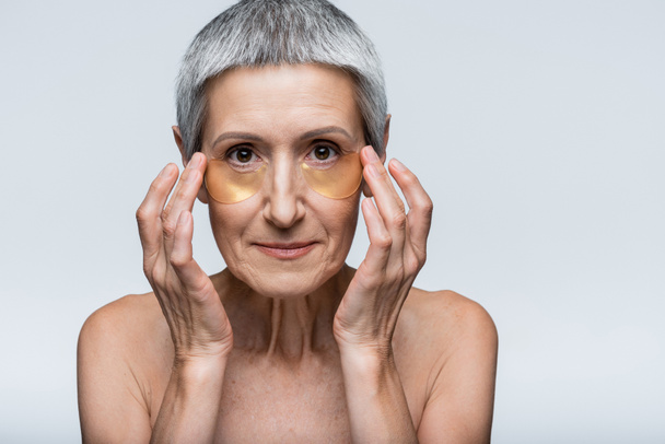 mujer de mediana edad aplicando parches de ojos dorados y mirando a la cámara aislada en gris - Foto, Imagen