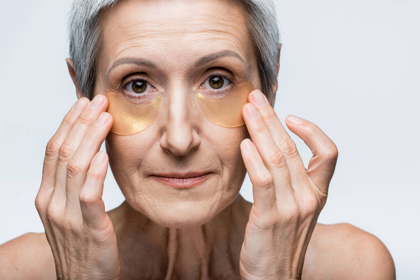 middle aged woman applying hydrogel eye patches and looking at camera isolated on grey - Foto, immagini
