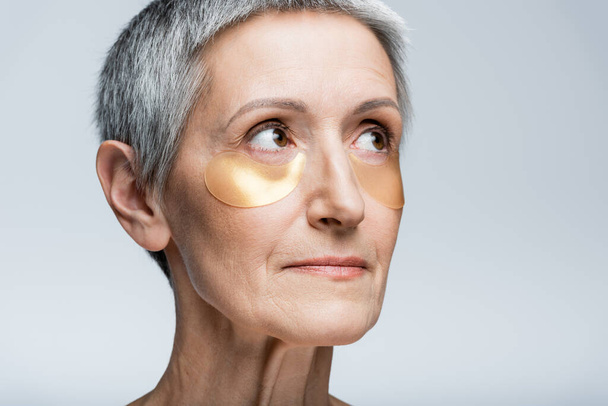 vrouw van middelbare leeftijd in gouden ooglapjes weg te kijken geïsoleerd op grijs - Foto, afbeelding
