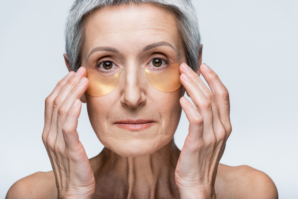 Frau mittleren Alters mit nackten Schultern, die goldene Augenflecken isoliert auf grau auftragen - Foto, Bild