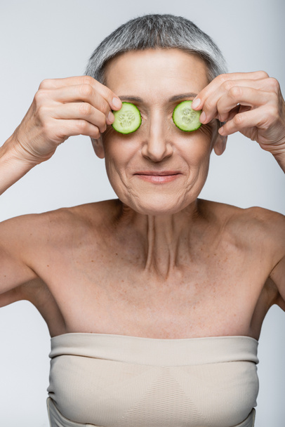 sorridente donna di mezza età che copre gli occhi con cetrioli tagliati verdi isolati su grigio - Foto, immagini