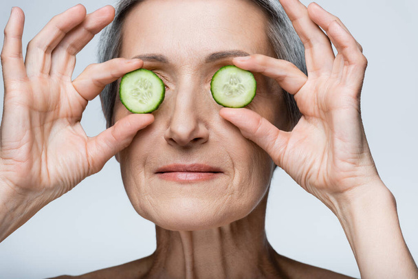 Nahaufnahme einer lächelnden Frau mittleren Alters, die Augen mit grünen Gurkenscheiben bedeckt, isoliert auf grau - Foto, Bild