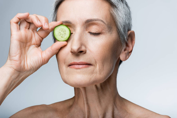 zralá žena zakrývající oko se zelenou nakrájenou okurkou izolovanou na šedé - Fotografie, Obrázek