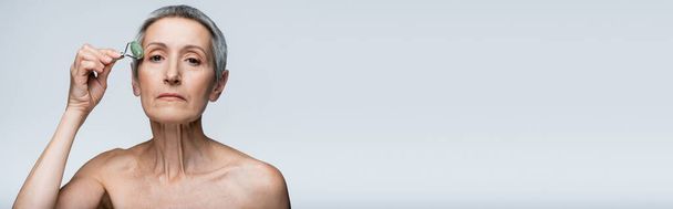Женщина средних лет с голыми плечами массирующее лицо с нефритовым роликом изолированы на сером, баннер - Фото, изображение