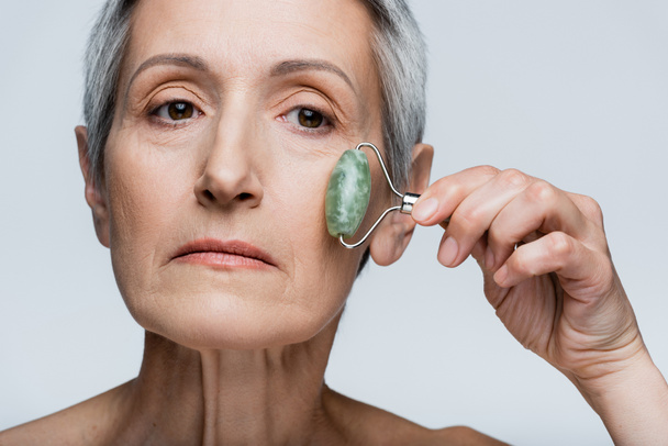 close up vista da mulher massageando bochecha com rolo de jade isolado em cinza  - Foto, Imagem