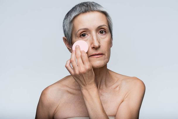 kobieta w średnim wieku stosując makijaż fundament z gąbki urody izolowane na szarości  - Zdjęcie, obraz