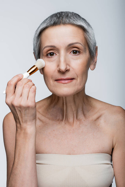 mature woman with grey hair applying face powder with cosmetic brush isolated on grey  - Foto, immagini