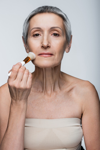 middle aged woman with grey hair applying face powder with cosmetic brush isolated on grey  - Foto, Imagen