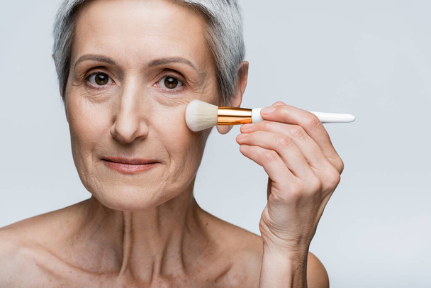 middle aged woman applying face powder isolated on grey - Φωτογραφία, εικόνα
