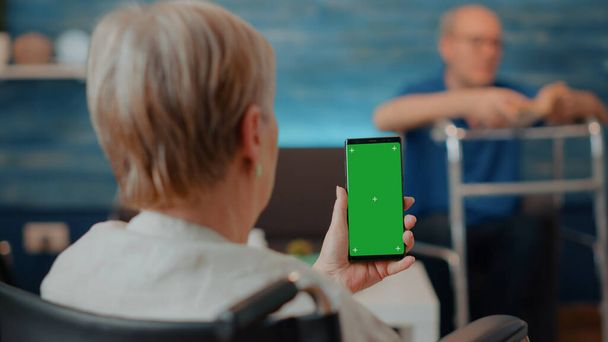 Tekerlekli sandalyedeki yaşlı kadın elinde yeşil ekranlı akıllı telefon tutuyor. - Fotoğraf, Görsel