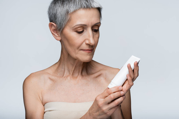 vrouw van middelbare leeftijd met grijs haar op zoek naar buis met lotion geïsoleerd op grijs  - Foto, afbeelding