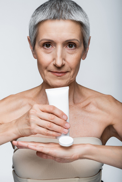 middle aged woman with grey hair holding tube and applying lotion on hand isolated on grey  - Foto, Imagem