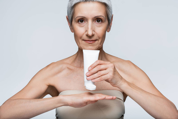 Frau mittleren Alters hält Schlauch und Anwendung Lotion auf der Hand isoliert auf grau  - Foto, Bild