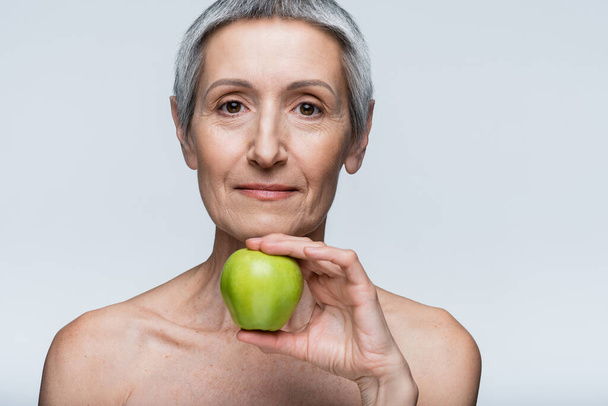Женщина средних лет с голыми плечами, держащая зеленое яблоко, изолированное на сером - Фото, изображение