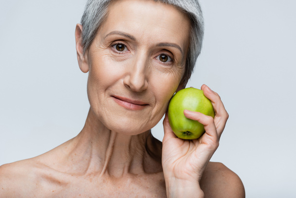 веселая женщина средних лет держит зеленое яблоко изолированы на сером - Фото, изображение