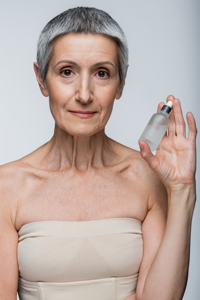 middle aged woman with grey hair holding bottle with serum isolated on grey - Foto, Imagem