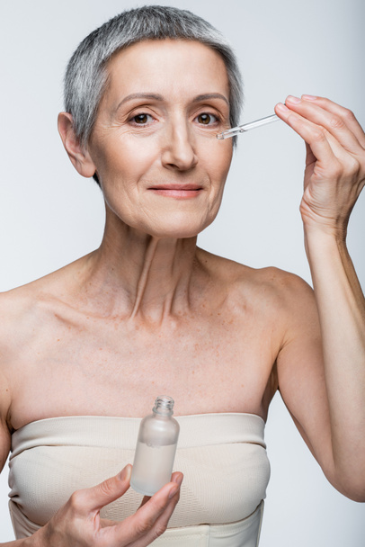 smiling middle aged woman holding bottle and applying serum isolated on grey - Fotó, kép