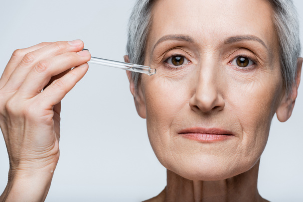 close up of middle aged woman with grey hair applying moisturizing serum isolated on grey - Fotografie, Obrázek