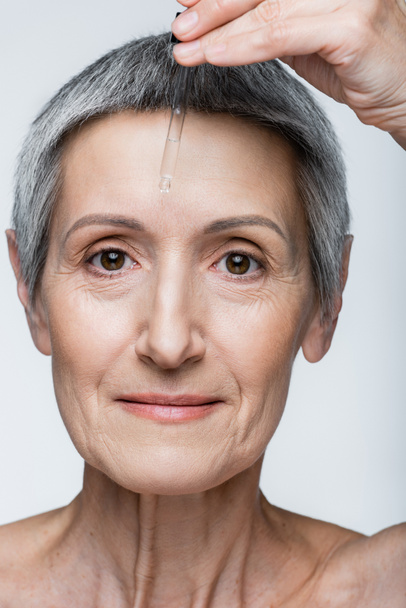 close up view of middle aged woman applying vitamin c serum isolated on grey - Foto, imagen